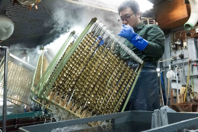 有限会社電研の工場内で、職人が金属部品を吊るしたラックを処理槽に浸す様子