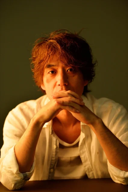 Hiro（川口博史）氏のポートレート、両手を組み顎に添えるポーズ