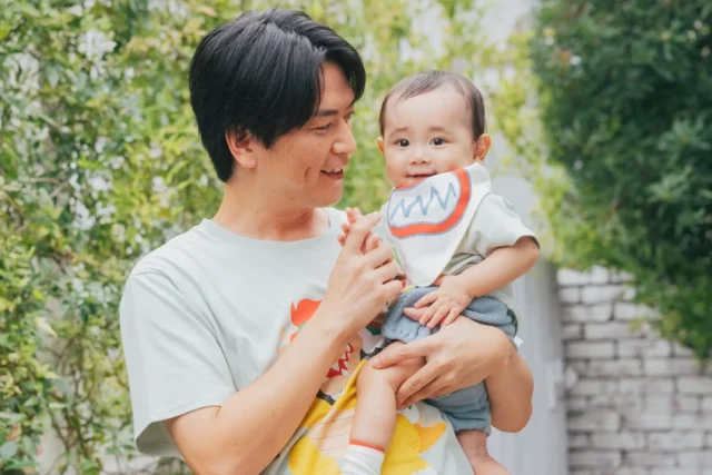 Tシャツ姿の男性が、よだれかけをつけた赤ちゃんを抱いている