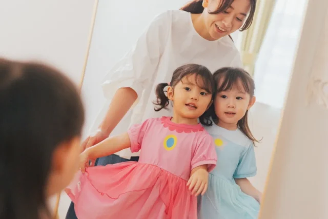 鏡の前でピンクと水色のワンピースを着た二人の子どもと女性