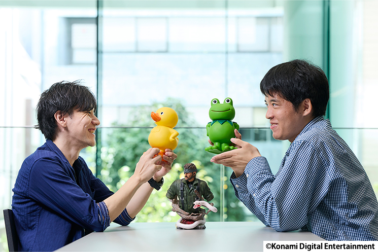 KONAMIのエンジニアが、ガーコとケロタンのフィギュアを手に持ち、談笑している様子